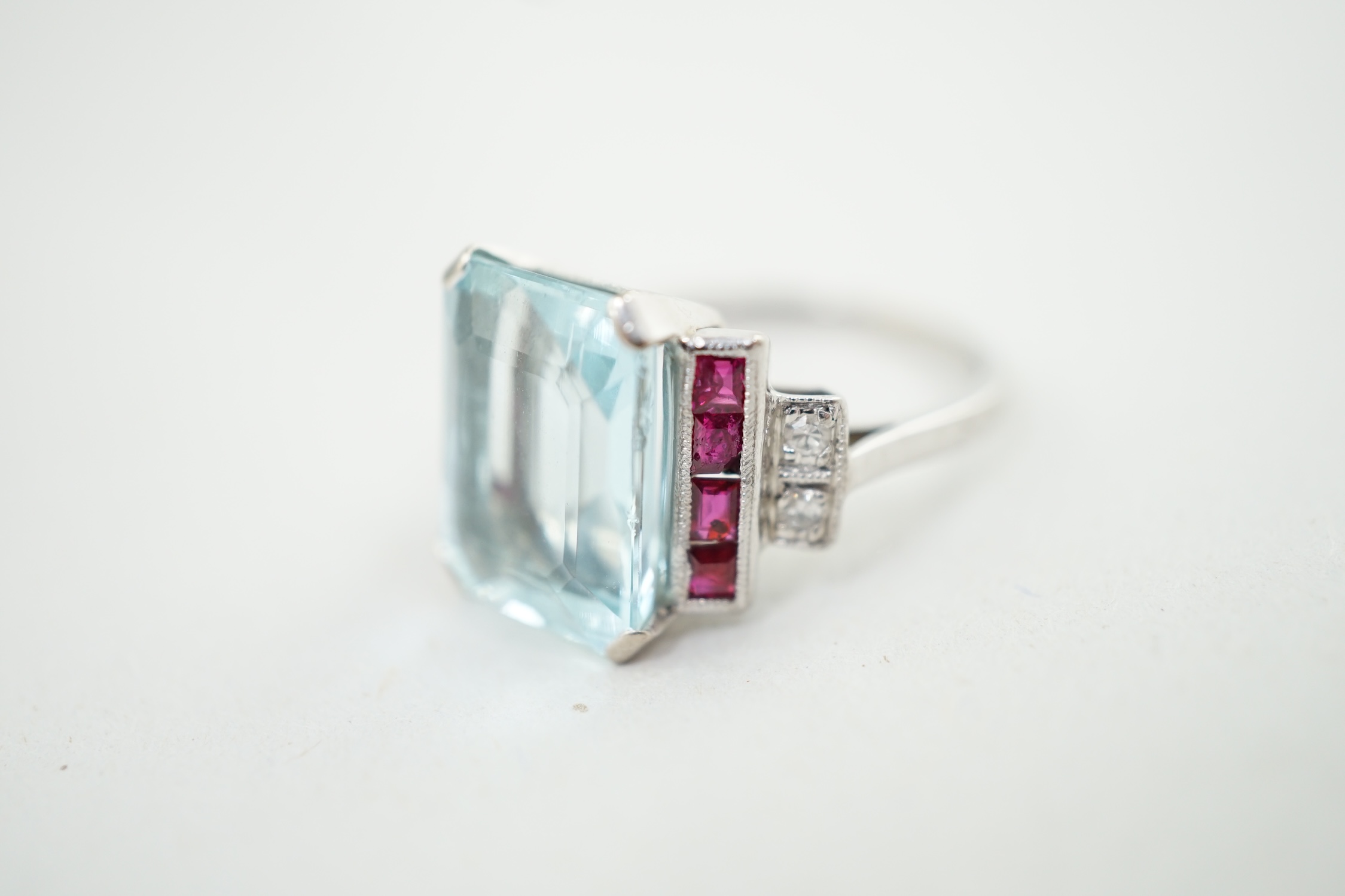 An Art Deco style platinum? and single stone emerald cut aquamarine set dress ring, with millegrain set ruby and diamond cluster stepped shoulders